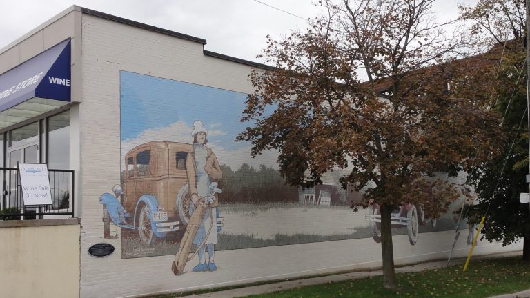 A mural painted on a white wall depicts a figure clad in 1920s golf attire, holding a set of golf clubs, standing in front of a 1920s-style car. The rest of the mural is obscured by a tree.