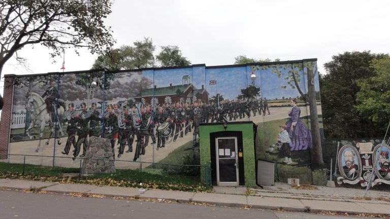 A mural painted on a wall depicts a military-dressed figure riding a horse in front of numerous military-clad figures playing and carrying instruments. Walking in rows, they parade in front of a school house and two watching figures, the smallest holding a drum. Two portraits, one labelled "Capt. Norris" is visible on the far right of the mural.