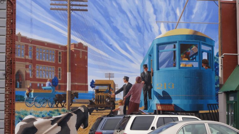 A mural depicting a blue tram trolley reading "213" and "Kingson" boarding passengers on an unpaved road. An early 20th-century vehicle waits beside the tram while a horse-drawn carriage rides in the opposing direction. A cow can be seen in the front left corner and a large building appears in the centre-left.