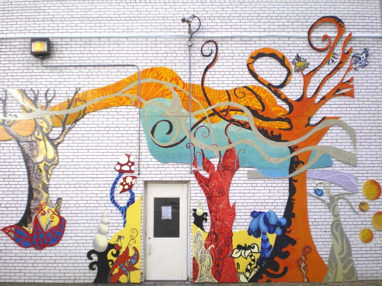 A mural with whimsically painted multicoloured trees, wind and fantasy fauna affixed to a white wall, surrounding a white utility door.