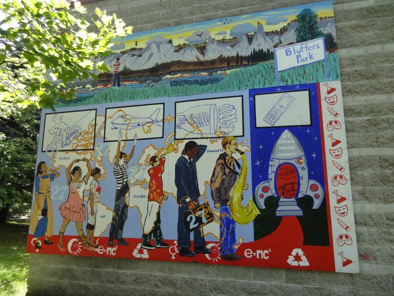 A mural affixed to a wall depicting seven figures demonstrating different artistic, healthcare and business professions stands in front of a rocket ship with the words "art," "science," "culture," and "community" written on the nose cone. A map of the world is painted as their background. Four images float above them, displaying a cyclist, an aeroplane, a lightbulb and a cellphone. A separate panel above shows an area with a sign labelled "Bluffers Park." A figure stands in the area, looking to see a cliff-face on a sunny day.