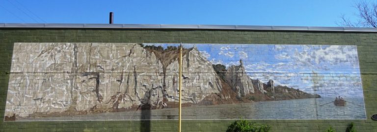 A mural painted on a side of a wall depicts a vast, grey cliffside beside a body of water on a sunny day. Visible in the water in a rowboat.