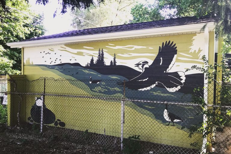 A mural painted on a wall depicts a bird soaring over a river and landscape. A frog is visible in the lower left corner, while a flock of birds fly in the upper left corner. Two Canada geese, one swimming in the water, one walking along the shoreline, are also visible.