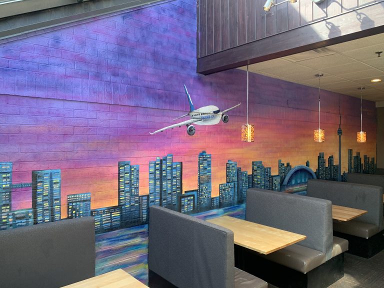 A mural of Toronto's skyline behind with a plane flying as the sun sets. Displayed behind a row of restaurant booths.