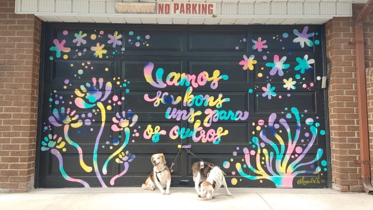 A black and rainbow coloured mural with the words 'Vamos ser bons uns para os outros' a phrase in Portguese meaning 'Let's be good to each other', featuring organic images surrounding the quote.