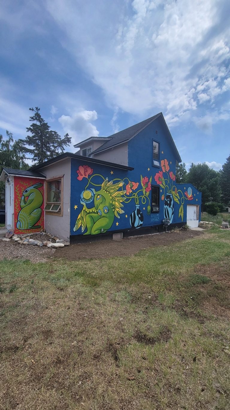 Painted house in vibrant colours of blue, green, pink, and yellow. Depicting a feminine mythical creature sitting with vases of poppies.
