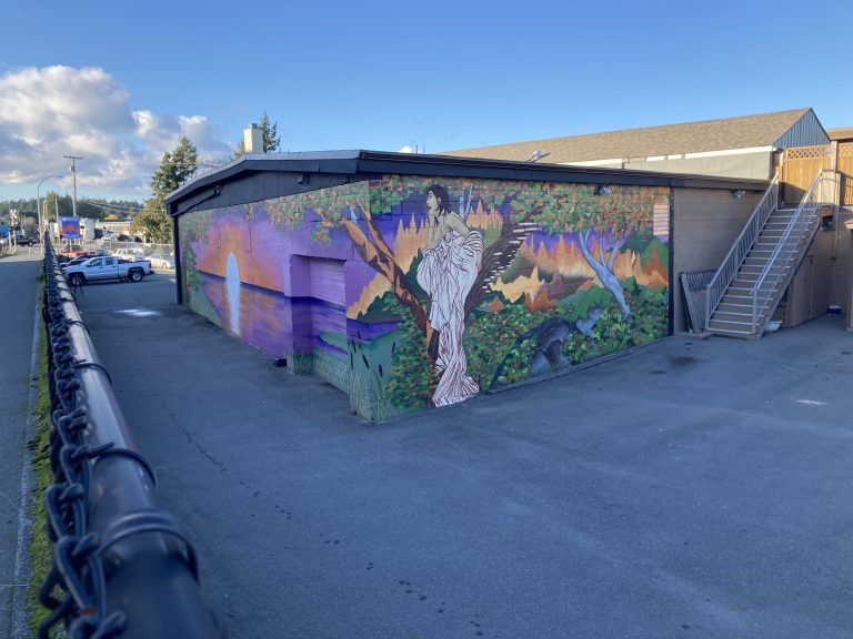 A mural painted on the corner or two walls of a warehouse, whose focal point is a figure in a long, flowing white dress, on one wall, who overlooks a sunset painted on the other wall. The woman sits in a tree, surrounded by an autumnal forest.