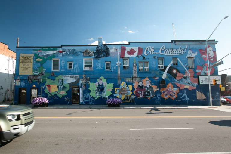 Photo of a large scale mural on the side of a building. Features a blue background with a Canadian flag and CN Tower at centre, and the text, “Oh… Canada!”. Around are various notable figures in Canadian history, each on a maple leaf background.