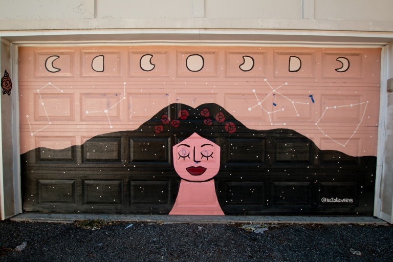Garage door mural of a female figure with hair extending across dotting with stars, with constellations and moon phases above her