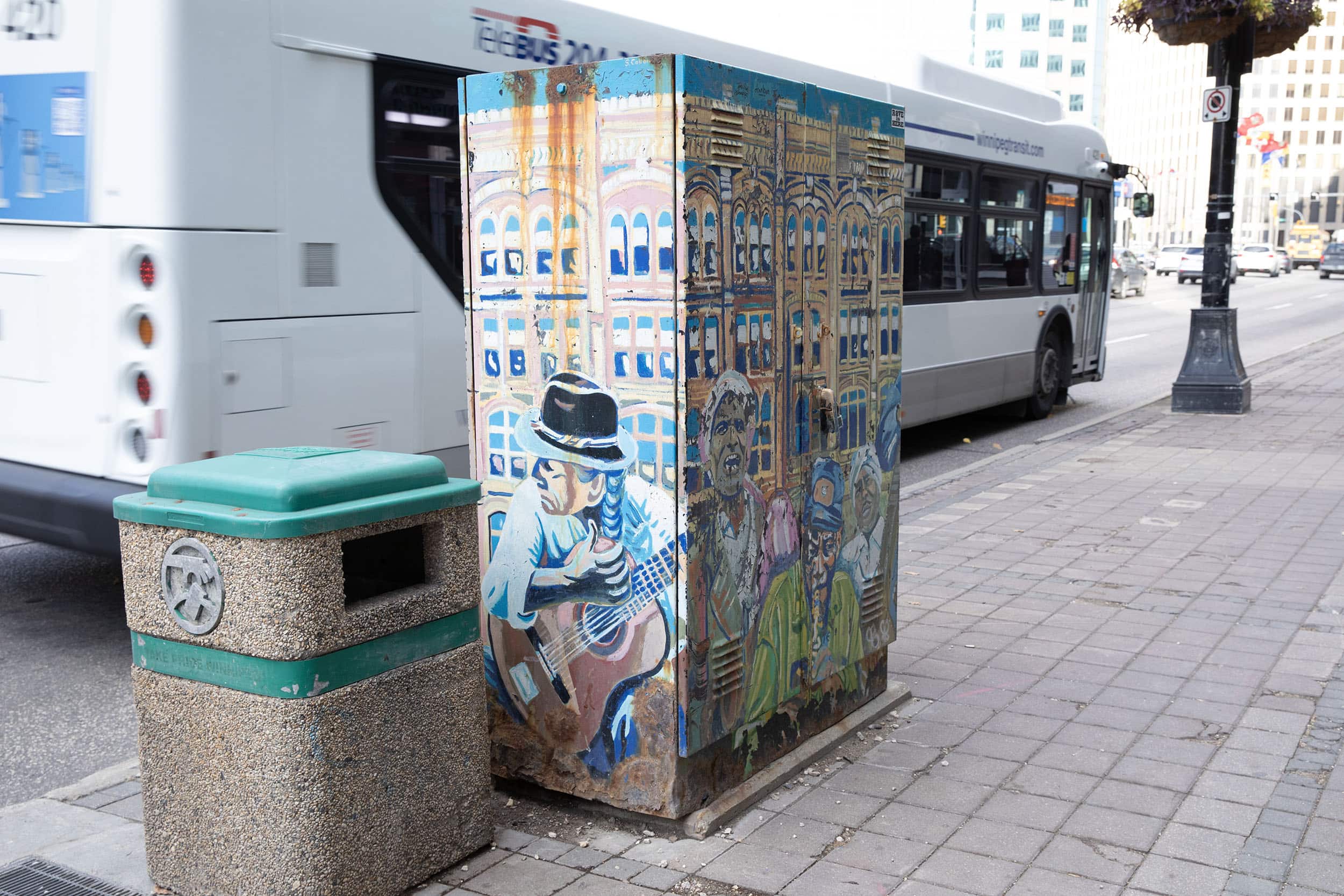 A transit box painting with bright, pastel colours depicts musicians playing instruments such as the guitar, saxophone, and violin. Others stand around these musicians laughing and smiling. The background of this painting is a neo-classical style stone building. Much of the paint has been chipped on this box. A city bus and distant traffic is seen passing behind the box.