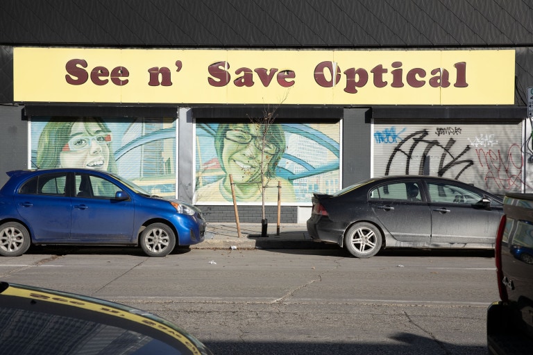 Two shutter panels, depicting a girl with red and blue glasses on one, and a boy with black glasses on the other. They stand inside a bus shelter, with a view of downtown Winnipeg behind them. The sign above these panels read "See n' Save Optical."