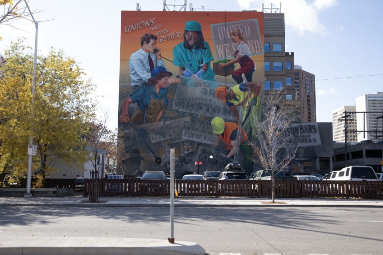 Images of various different workers on an organe background. Text at the top reads "Unions Make Work Better." In the distance, a crowd of strikers are seen in black and white. They hold signs that read "United we stand," "We asked for bread and recieved a stone," "One for all, all-for one," and "We fight for the right to a living wage."