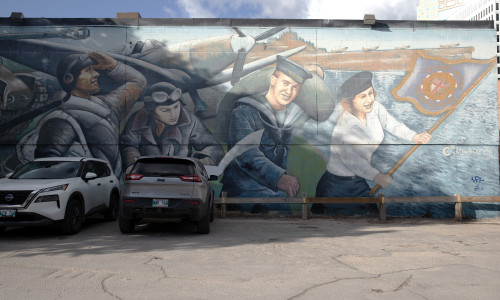 Four of six figures are visible in this photo of the mural. These figures stand in a line behind an army plane and the sea on the right side. They consist of three pilots, and a naval wren who holds a flag.