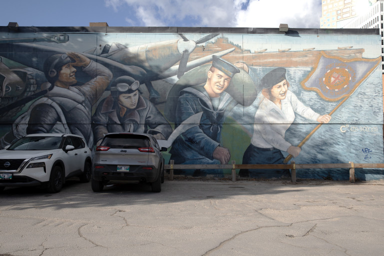 Four of six figures are visible in this photo of the mural. These figures stand in a line behind an army plane and the sea on the right side. They consist of three pilots, and a naval wren who holds a flag.