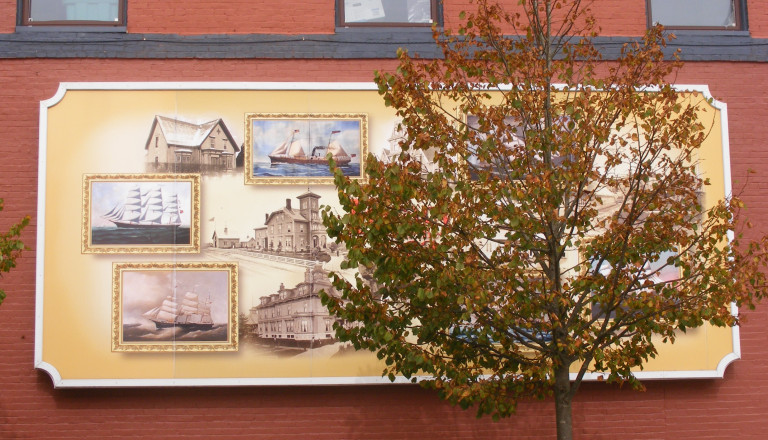 A wall features a framed mural, somewhat covered by the leaves of a tree. The mural contains snapshots of historic houses and boats.