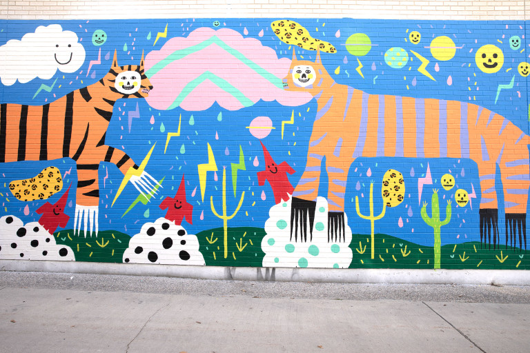 Brightly painted mural on a brick wall, depicting two tigers who face eachother. They have long claws and white masks on their face. They stand in front of a blue background with dark green grass, with white clouds near the bottom, with red figures coming out of them. The background is filled with bright colours, shapes, and patterns succh as clouds, lightning bolds, planets, smiley faces and confetti.