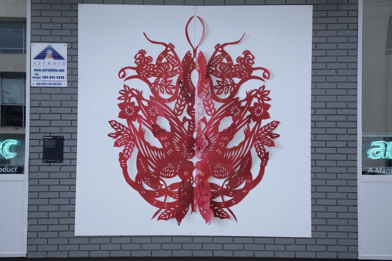 Photograph of a red paper cut artpiece. Shapes of leaves, flowers and parts of animals are seen and mirrored vertically.
