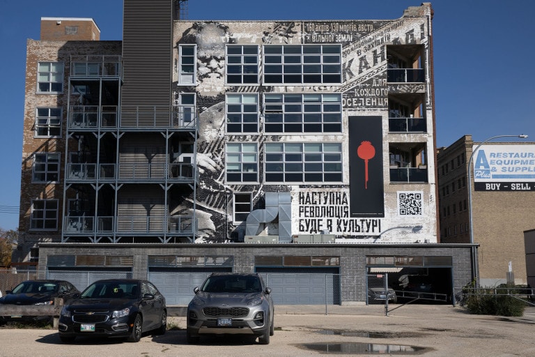 A large warehouse building with many windows is painted with words in Ukranian, a male figure is painted in between windows on the left side. On the right side, is a long panel with an image of a poppy.