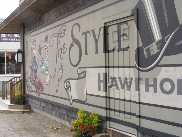 The side of a building is painted sage green with characters and objects drawn in 1920s style: a woman with a feathered hat, perfume bottles, a mirror and a woman in a corset sitting on a carriage pulled by lobster.