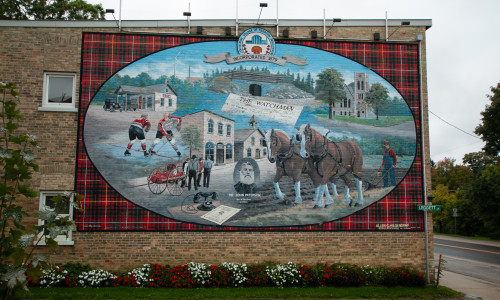 Image of a large heritage mural on the side of the building with subject of hockey players, carriages and other landscape features of the area in an oval shape, over a red plaid background.