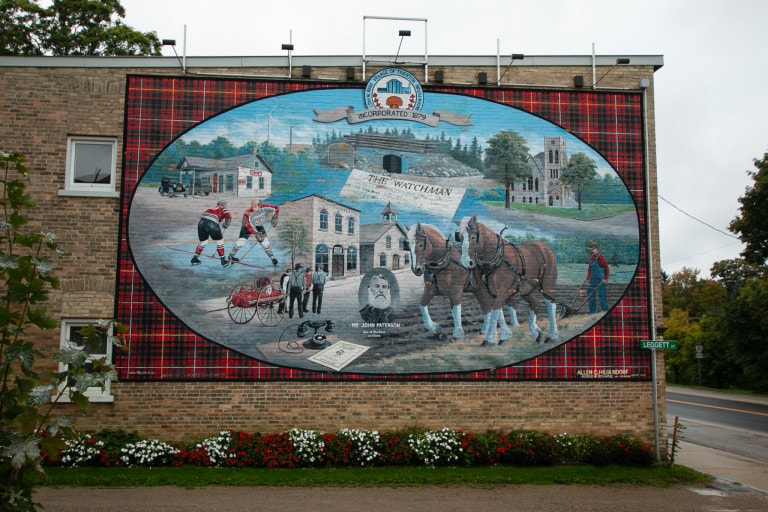 Image of a large heritage mural on the side of the building with subject of hockey players, carriages and other landscape features of the area in an oval shape, over a red plaid background.