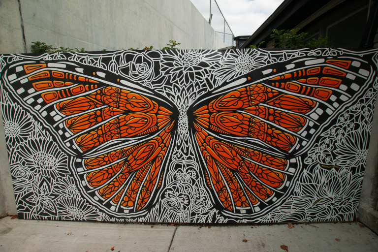 Photo of a mural on panel of a monarch butterfly in decorative patterned linework on the wings. The background is intricate linework of various flowers in white.