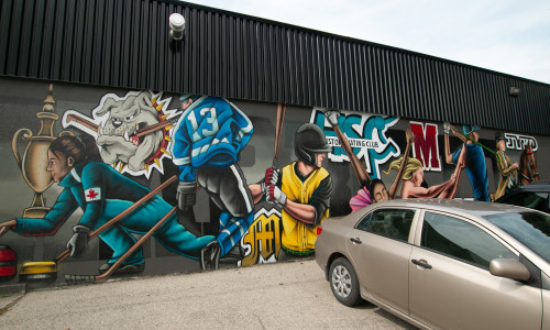 Photo of a mural by a parked car. Mural shows a collage of different sports players.