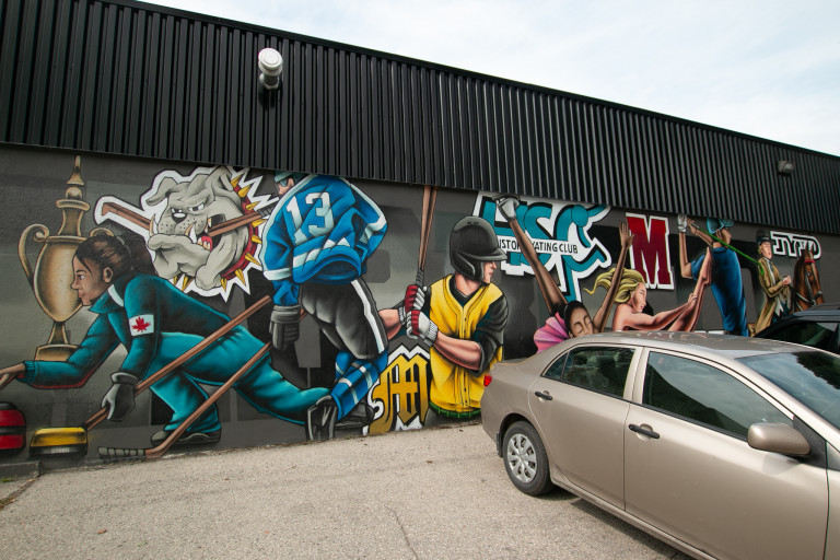 Photo of a mural by a parked car. Mural shows a collage of different sports players.