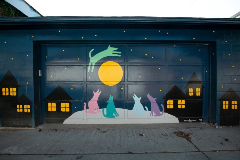 A garage door mural of a night scene with black houses set against a dark blue background. The houses have yellow windows suggesting that the lights are on. In the centre of the image is a green dog jumping over a large yellow moon. Below the dog and moon are 4 more dogs looking up from a giant rock or snow bank. The dogs looking up are pink, teal, light blue and purple. There are yellow stars dotted throughout the sky.