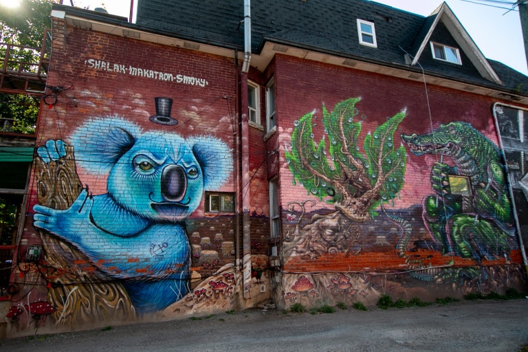 Mural on side of building with an angry blue koala with a mustache, on the left of a rainforest tree house, with a large crocodile beside the tree.