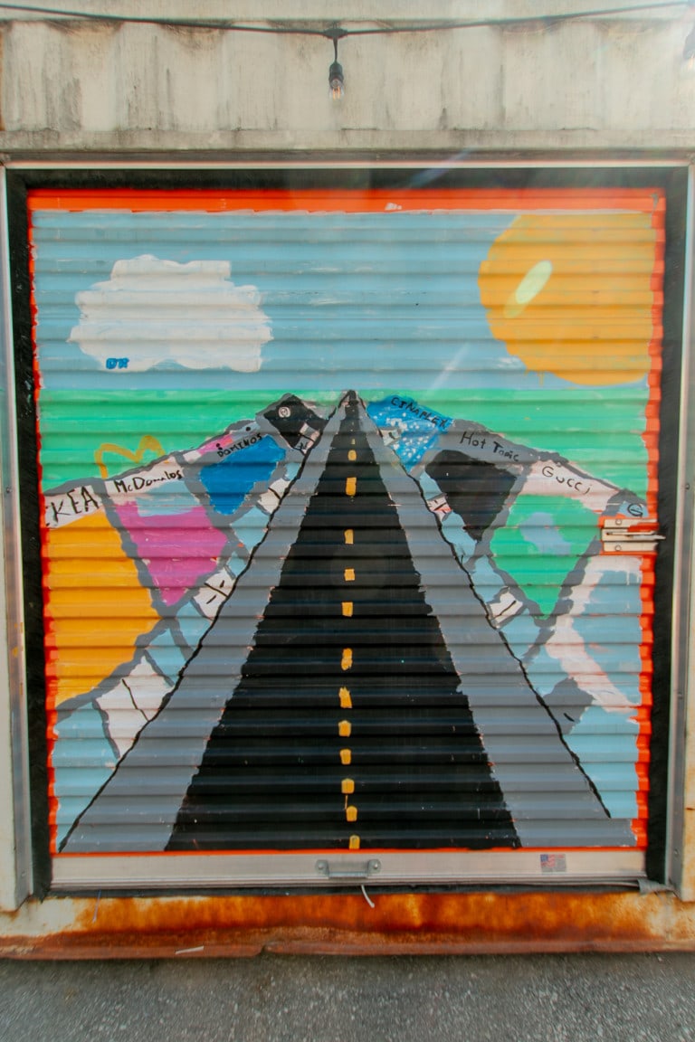 Image of a mural painted by youth of a black road with yellow divisors going into the horizon of a blue sky and a large sun on the right. On the road sides are familiar stores. On left, the stores are labeled: IKEA, McDonald’s, Dominos and KFC. On the right: Cineplex, Hot Topic, Gucci.
