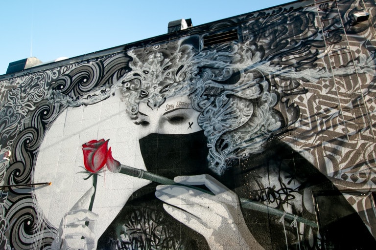 Image of black and white mural featuring a woman wearing a mask wearing a headdress. She is painting a rose which is coloured red, the only colour seen in the piece. Around her are symbolic motifs set in a circle.