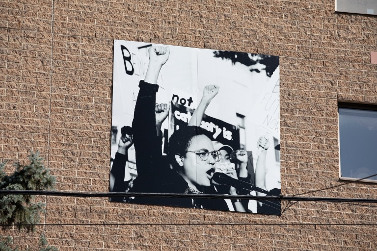 Photo panel located on a brick wall. The photo is a black and white image of a person with glasses, with their fist in the air, speaking into a microphone. Behind them, others raise their fists and hold rally signs.