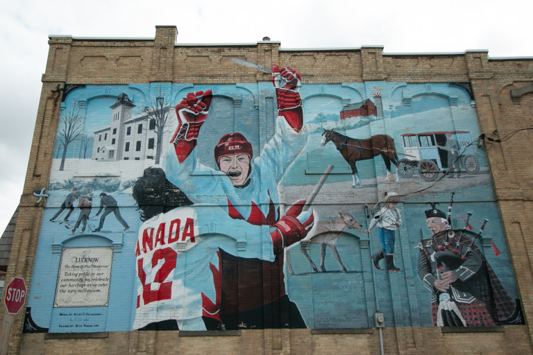 Photo of mural of a victorious hockey player with his arms up embraced by another player, background features a horse and carriage, people playing hockey outdoors, a man leading a deer, and a man with bagpipes.