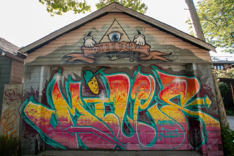 Garage door mural of a graffiti in red, yellow and purple of “Miles”. A triangle character with a single eye floats above it, holding a banner that says “Miles” in a typography font.