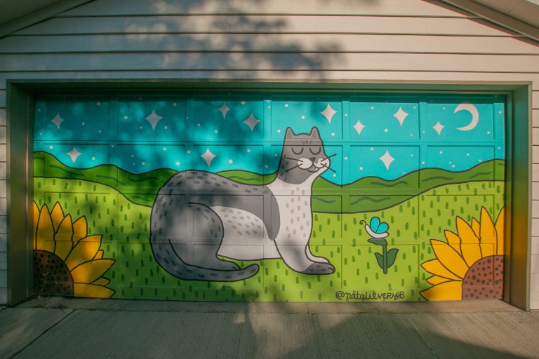 Garage door mural of a smiling cartoon grey and white cat on a green field with a light blue sky dotted with stars and featuring a crescent moon on the top right. Two sunflowers are at the foreground at the bottom left and right corners.