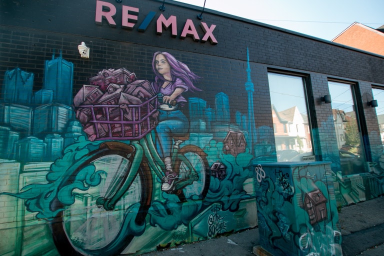 Mural for Remax Ultimate in aerosol, of a woman riding a bike in the Toronto city scape, with her bike basket full of houses. Background is black, and main hue of background is blue. The woman is sporting purple hair and wearing a purple shirt. The houses in the basket are also purple.
