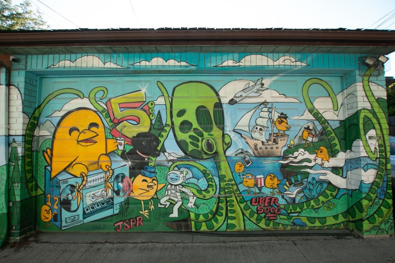 Garage door mural of a chick on a boom box in a day time scene with an octopus at the forefront with a dog and a spaceman, and a ship in the background on the water.
