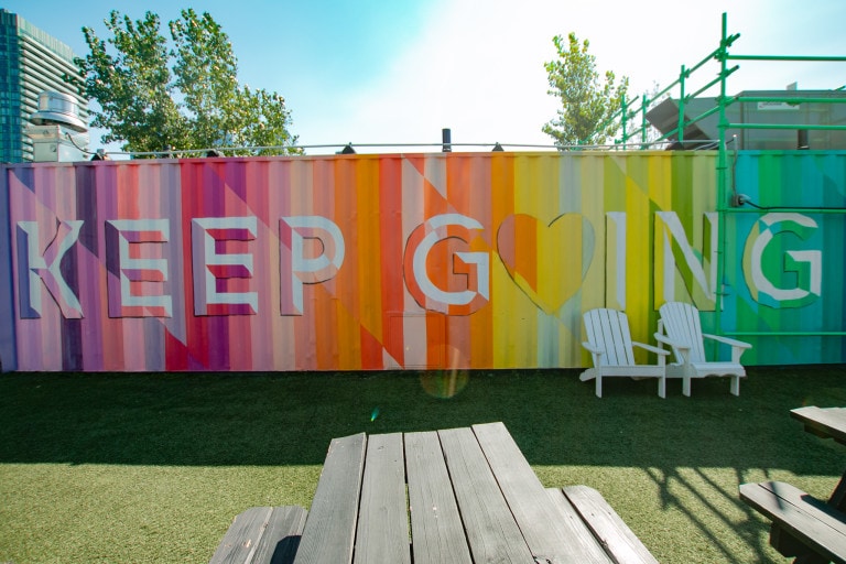 Colourful mural on a shipping container with lined vertical gradients in a glitch-like rainbow colour, featuring text in block letters, “Keep Going.”