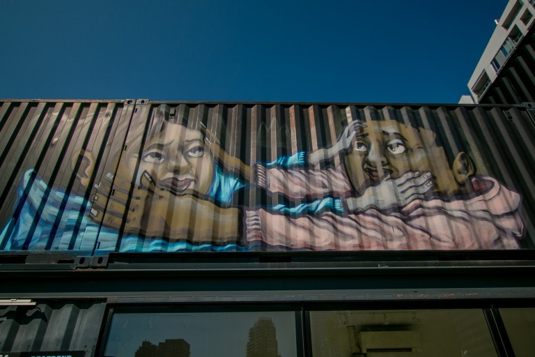 Photo of a shipping container mural from ground level of two characters touching each other’s faces in what appears to be affection. One is a female with a light blue hoodie, and the other is a male with a peach hoodie.