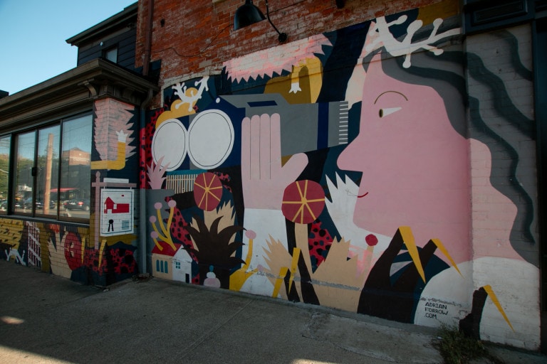 Mural on side of building of a pink lady with her hand up, smiling and looking out to a number of patterns and graphics in red and yellow tones. Background is navy blue
