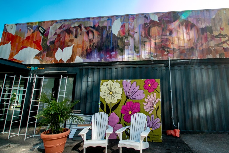 Photo of a shipping container mural above a store featuring two portraits in abstract realism. A man and woman are laying down with their heads towards each other, body is not shown. The man is vaguely smiling and his arm is seen, perhaps holding a phone. The woman has a relaxed face. Abstract motifs cover the mural in dreamy oranges and purples, along with white leaf-shaped blocks.