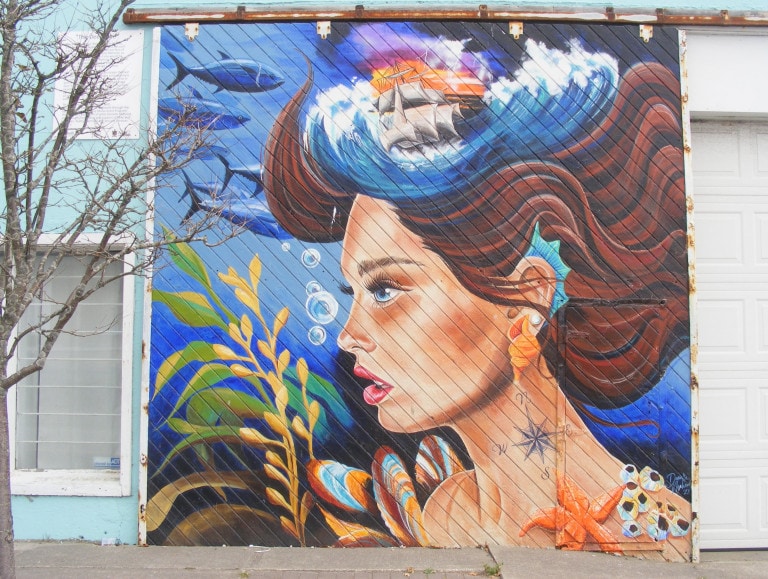 A portrait of a young woman underwater is painted on the side of a local business. She is surround by ocean vegetation, shells, different kinds of fish, a starfish and bubbles. A sailboat and waves are perched atop her head.