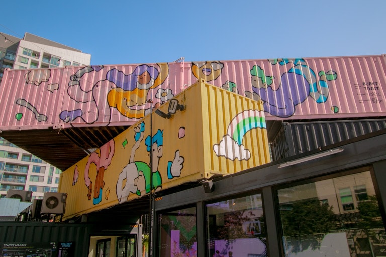 Photo of two shipping containers stacked on top of each other above a store, each with a mural painted by the same artist. The top mural is in pink and features two floating cartoon characters wearing orange and purple, respectively. The shipping container below is painted in yellow and features two other characters, one i upside down and appears to be waving, the other is holding what appears to be a phone. Around the characters are various objects including a spoon, a heart emoji face, and a thumbs up emoji.