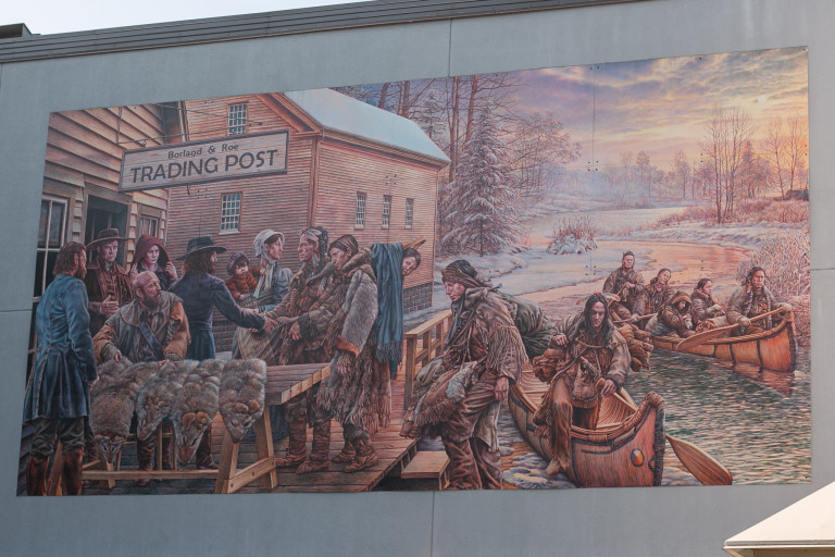 Photo of a realistic mural in brown tones featuring people at a trading post, with a sign writing “Borland & Roe Trading Post.” Fur pets are seen hanging over a bench with what appears to be European settlers, and indigenous people are on the right coming up from canoes.