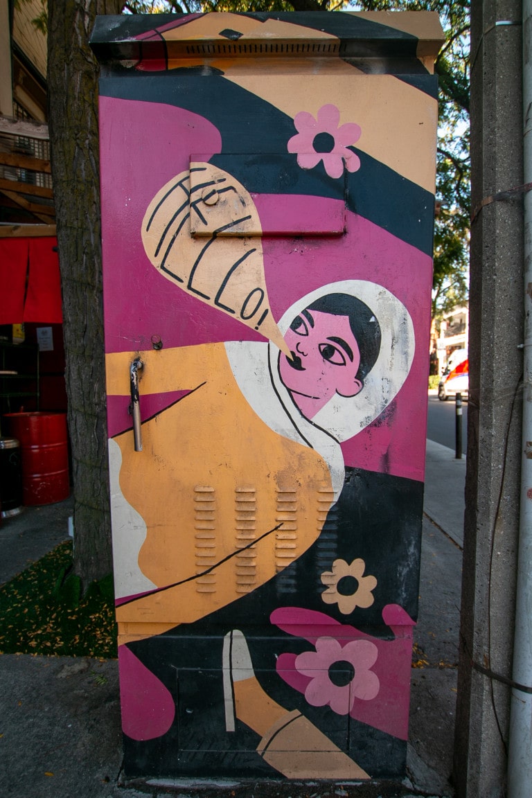 Illustrative painting on a traffic box of a floating figure in a space suit saying “Hello!” Background is pink and black with flowers, figure is in yellow