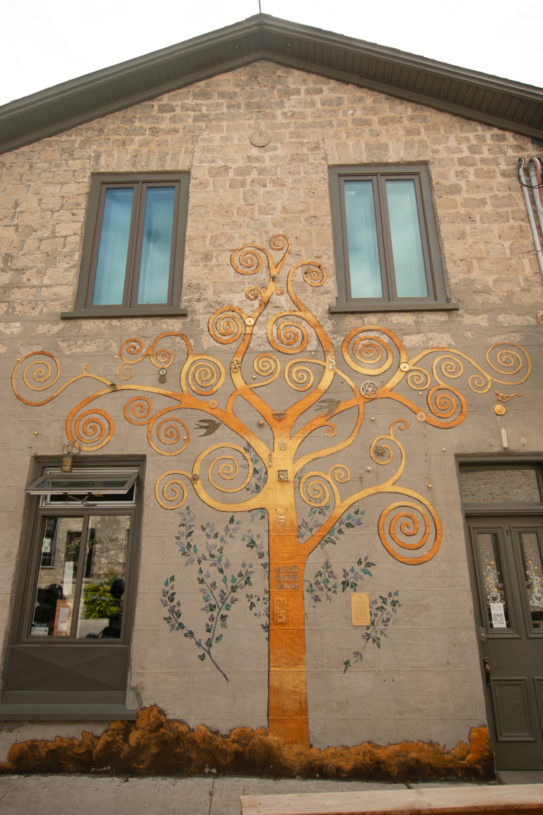 Photo of a mural on a brick house of the tree of life in guilded brass. The tree is thin with branches swirling out, and 3D found objects are seen along the tree trunk.