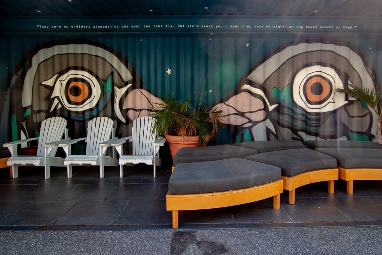 Photo of an aerosol mural behind chairs. Mural is on corrugation and has a dark blue and green background remniscient of the northern lights. The subject features two pigeon heads facing each other in a linework style.