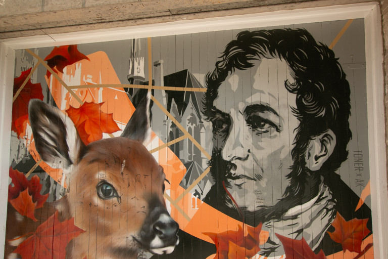 Photo of a mural on wood of a man in black and white and a deer at the left forefront in colour, with maple leaves around it.