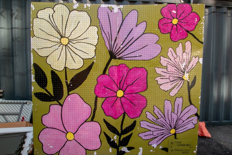 Image of a mural on a metal board with a yellow-green background and various large flowers painted in pinks and purples. One white daisy-like flower sits at the top left.